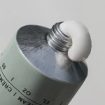 Close-up of a hand cream tube dispensing white cream from its metallic nozzle.