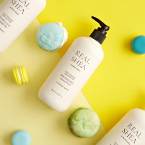 White shampoo bottle labeled Real Shea on a bright yellow background with pastel macarons nearby.