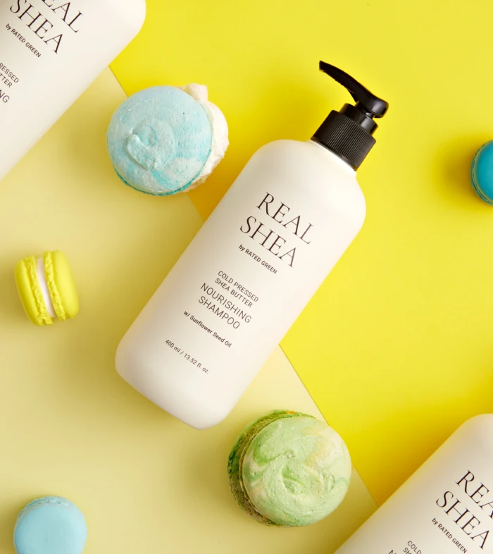 White shampoo bottle labeled Real Shea on a bright yellow background with pastel macarons nearby.