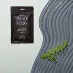 Black sachet labeled 'ROSE MARY' with 'BALANCING SCALP PACK' and 'w/ Charcoal' beside a textured gray surface and green plant fronds on the right.