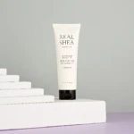 White skincare tube labeled Real Shea by Rated Green standing upright on white stacked steps against a gray studio backdrop