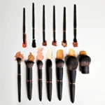 Row of black makeup brushes lined up with colored powder swatches beneath each brush on a white background, branding visible on handles.