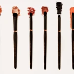 Seven makeup brushes lined up vertically with crushed red, pink, and black pigment on the bristles against a white background, showcasing makeup powders.