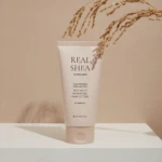 White skincare tube labeled REAL SHEA with dried wheat stalks resting on/over it against a warm beige backdrop.