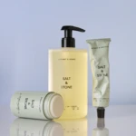Salt & Stone skincare set: pale yellow pump bottle with two mint-green tubes against a blue background, minimalist typography visible.