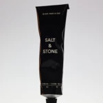 Glossy black squeeze tube standing upright against a light gradient background, with gold text reading 'SALT & STONE' on the front.