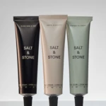 Three Salt & Stone hand cream tubes in black, beige, and sage green standing upright from left to right.