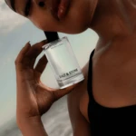 Close-up of a person holding a Salt & Stone perfume bottle near their lips against a pale sky background (Bergamot & Hinoki).