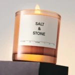 Lit pink glass candle with a small flame, label reads 'SALT & STONE' and scent note 'SAFFRON & CEDAR'.