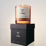 Amber glass candle with a white label reading 'SALT & STONE' perched on stacked black boxes against a pale background
