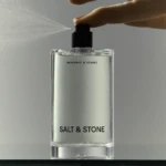 Close-up of a perfume bottle being sprayed, with mist spreading to the left; label reads Bergamot & Hinoki, Salt & Stone.