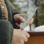 Person applying white cream from a light gray tube labeled 'Salt & Stone' onto the forearm outdoors, wearing a green fleece jacket.