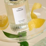 Close-up of a glass perfume bottle labeled 'Anillo Amber528' resting on a glass pedestal with lemon peels nearby.