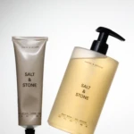 Two Santal & Vetiver Salt & Stone products: a beige lotion tube and a pale yellow pump bottle angled on a reflective surface with their reflections in the water.