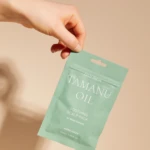 Hand holding a teal Tamanu oil sachet against a beige background, with 'Cold Press' and 'Tamanu Oil' text visible on the package.