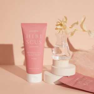 Pink tube of Hibiscus Moisturizing Scalp Pack in front of a peach background with a clear vase and dried flower nearby.