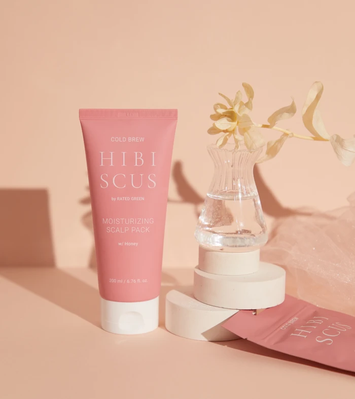Pink tube of Hibiscus Moisturizing Scalp Pack in front of a peach background with a clear vase and dried flower nearby.