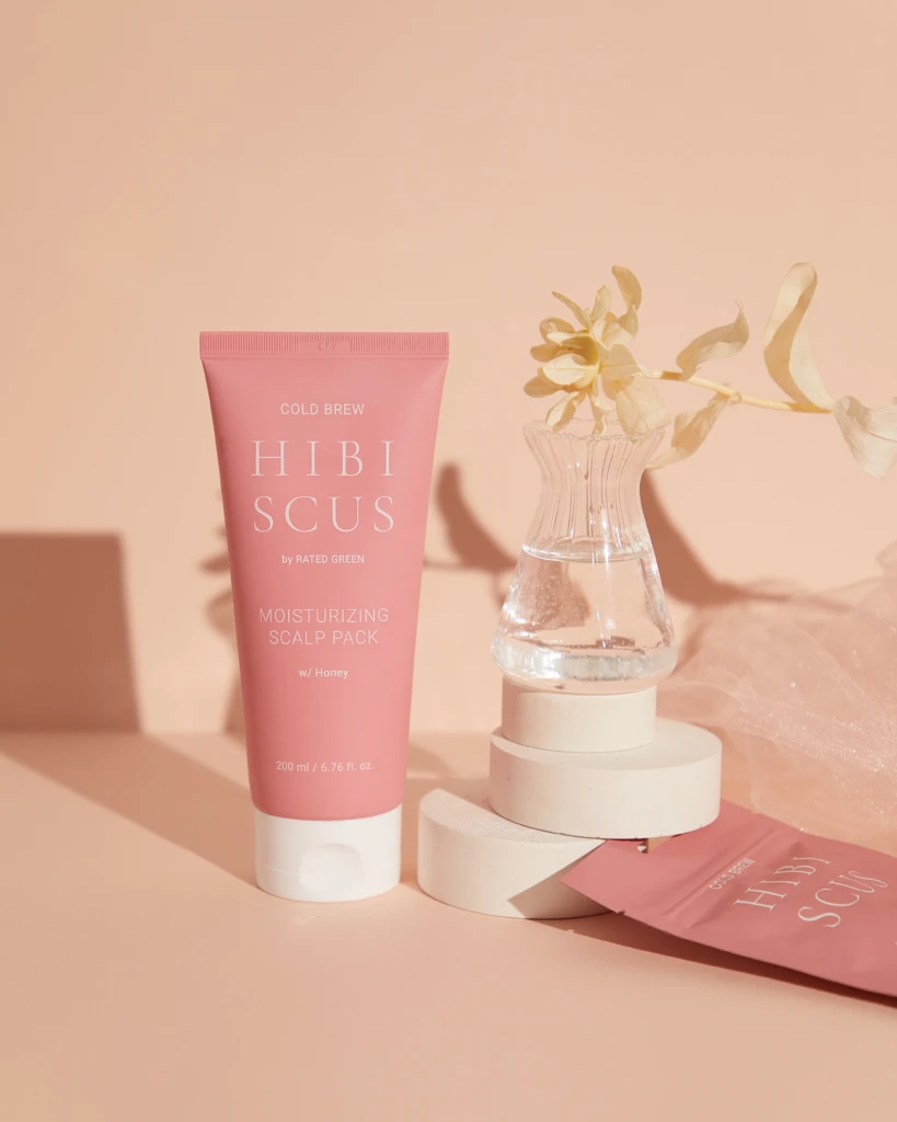 Pink tube of Hibiscus Moisturizing Scalp Pack in front of a peach background with a clear vase and dried flower nearby.