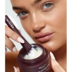 Close-up of a woman scooping white cream from a burgundy BB Skin Vitamin Hydration jar with a spatula.
