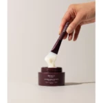Hand holding a spatula with white cream above an open dark purple jar labeled 'BBSKIN Vitamin Hydration Mask'.