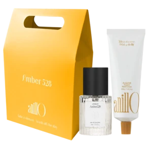 Bright yellow gift-box style package with a built-in handle, accompanied by a clear glass fragrance bottle labeled Amber528 and white boxed packaging nearby.