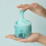 Teal body scrub in a clear jar being held with two hands, showcasing the product's creamy texture as it stretches from the lid opening. (Shower Time Body Scrub)