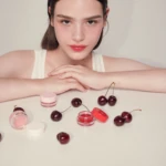 Young woman with subtle makeup sits at a white table with pink makeup jars and cherries scattered nearby.