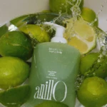 A glass bowl filled with green limes and a green shampoo bottle, with water splashing over the fruit and bottle.
