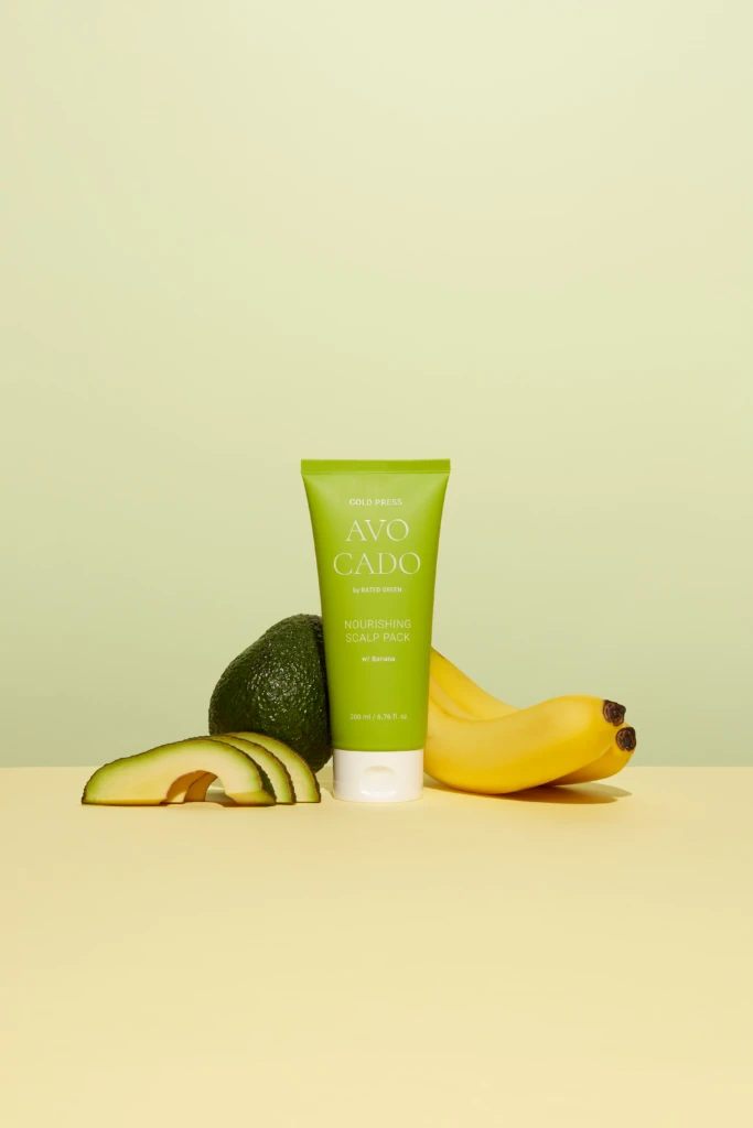Green tube of Avocado Nourishing Scalp Pack with avocado halves and bananas surrounding it against a pale yellow backdrop