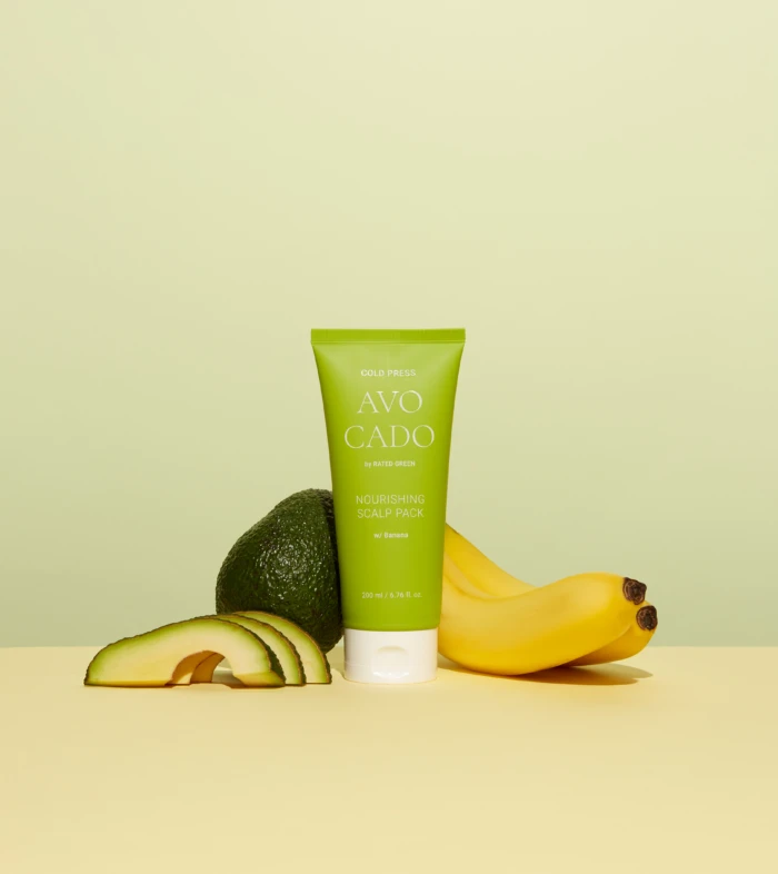 Green tube of Avocado Nourishing Scalp Pack with avocado halves and bananas surrounding it against a pale yellow backdrop