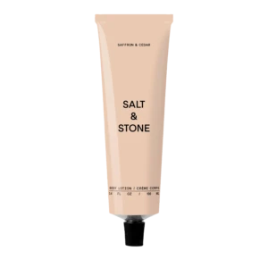 Pale pink body lotion tube standing upright; reads 'Salt & Stone' with 'Saffron & Cedar' scent on a black background.