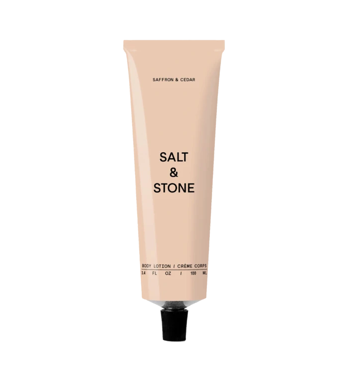 Pale pink body lotion tube standing upright; reads 'Salt & Stone' with 'Saffron & Cedar' scent on a black background.