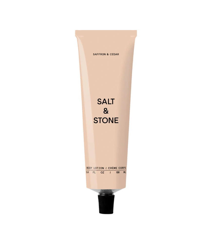 Pale pink body lotion tube standing upright; reads 'Salt & Stone' with 'Saffron & Cedar' scent on a black background.