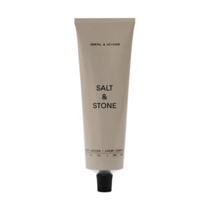 Beige squeeze tube labeled 'SALT & STONE' with a black cap, standing upright with 'Body Lotion' text at the bottom against a black background.