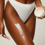 Person in white ribbed underwear applying white cream to their thigh from a jar held in one hand