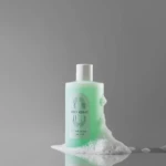 Mint-green bottle of Acne Derma Wash with white cap, surrounded by white foam on a reflective surface against a gray background.