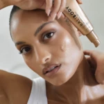 Close-up of a woman with dewy skin holding a rose-gold tube labeled illuminating serum near her temple.