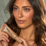 Close-up of a woman with wavy brown hair holding a beige tube labeled 'BABY' bronzing serum near her face.