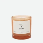 Brown glass candle jar with a white label reading 'SALT & STONE' on a light background, close-up shot, partial view of the top edge visible.
