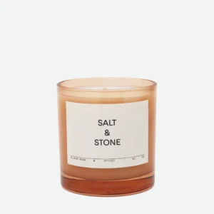 Brown glass candle jar with a white label reading 'SALT & STONE' on a light background, close-up shot, partial view of the top edge visible.