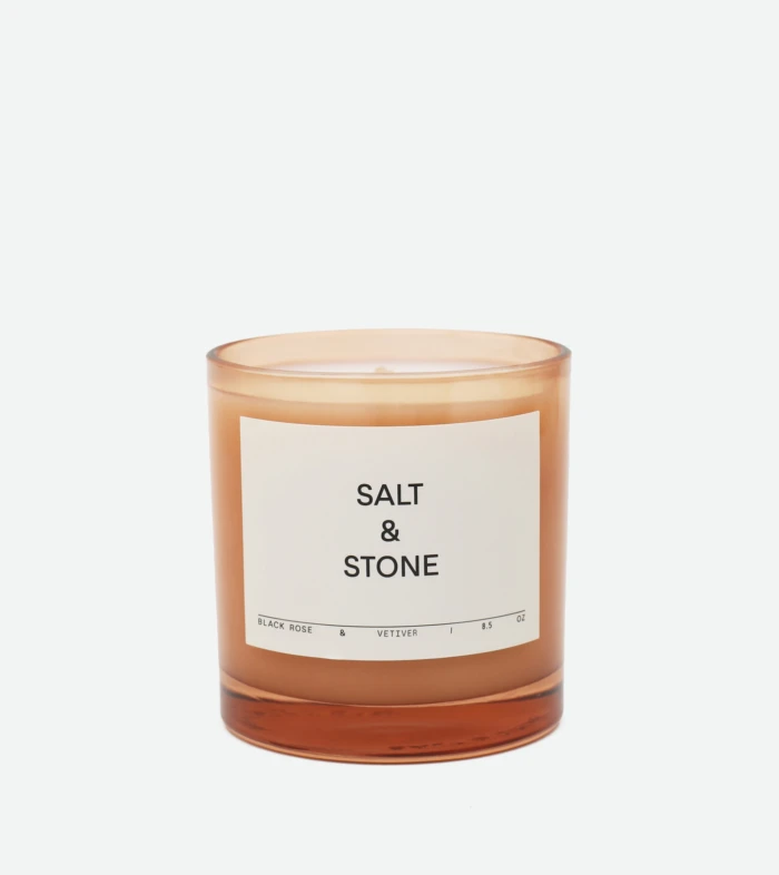 Brown glass candle jar with a white label reading 'SALT & STONE' on a light background, close-up shot, partial view of the top edge visible.
