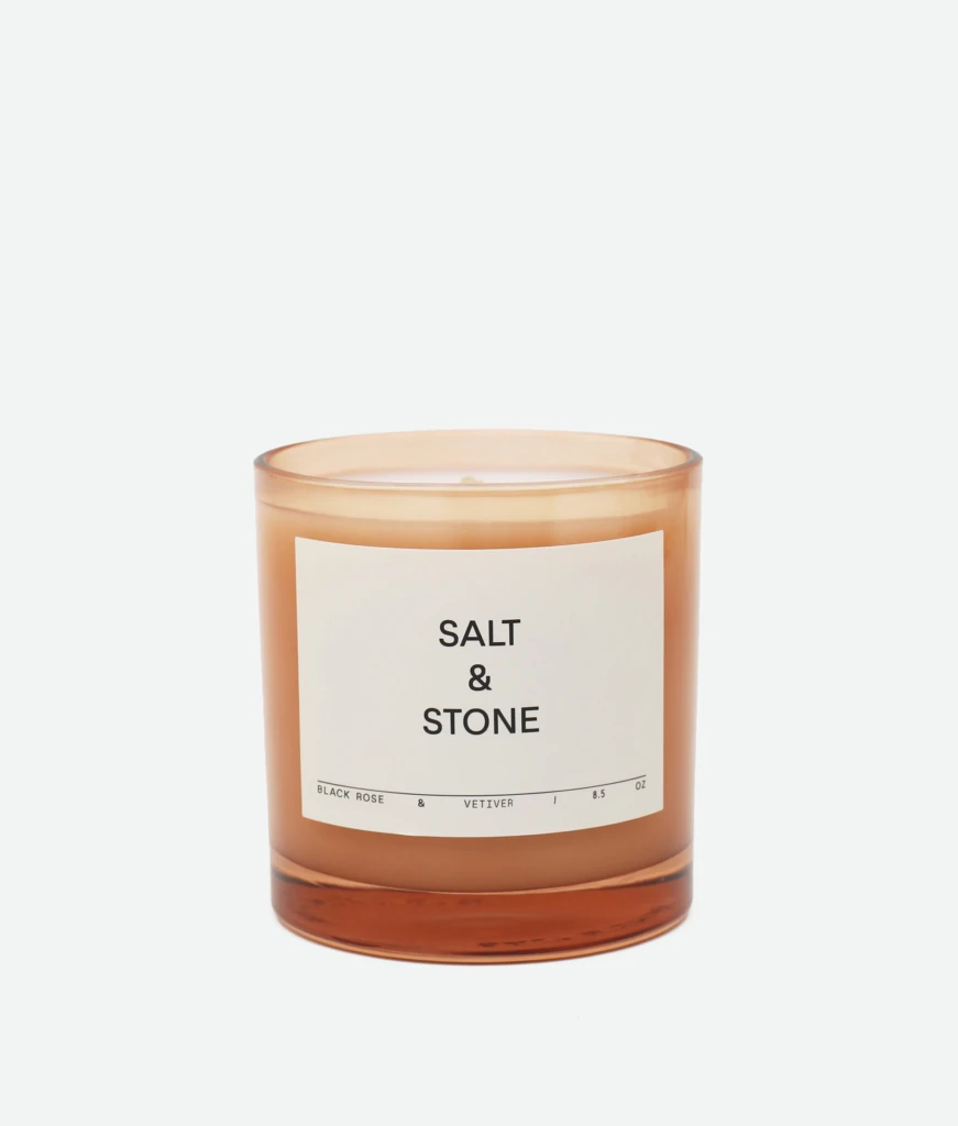 Brown glass candle jar with a white label reading 'SALT & STONE' on a light background, close-up shot, partial view of the top edge visible.