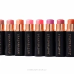 Row of black lipstick tubes with rose-gold caps in a line, showing pink to coral lipstick shades on a white background.