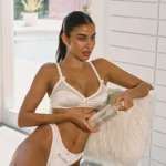 Woman in a white lingerie set sits by a white brick wall, looking to the side with a calm expression.