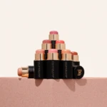Tower of lipstick tubes in pink and nude shades stacked in a pyramid on a light pink surface, showing black and gold packaging.
