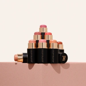 Tower of lipstick tubes in pink and nude shades stacked in a pyramid on a light pink surface, showing black and gold packaging.