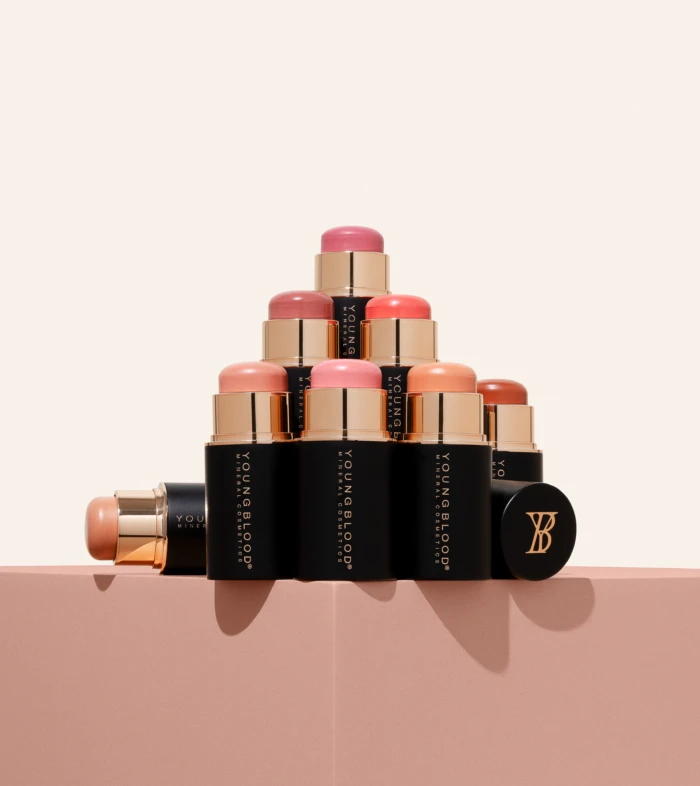 Tower of lipstick tubes in pink and nude shades stacked in a pyramid on a light pink surface, showing black and gold packaging.