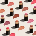 Row of black lipstick tubes with nude-pink swatches scattered across a pale background, showcasing shade range.