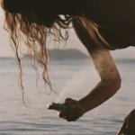Close-up of a person’s hand reaching toward the camera over water, with wet hair and a sunlit scene in the background.