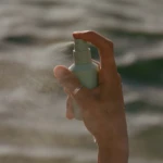 Close-up of a hand pressing the spray nozzle of a pale green bottle, releasing a fine mist outdoors.